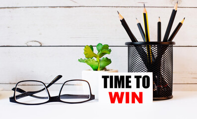 TIME TO WIN written on a white business card next to pencils in a stand and glasses. Nearby is a potted plant.