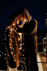 Fototapeta na wymiar Portrait of beautiful couple with light garland stand at the balcony, handsome man hug gorgeous woman, enjoy tender moments, winter holidays, Christmas celebration concept