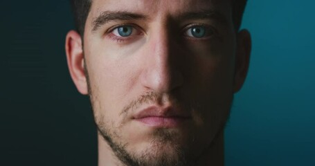 Cinematic close up of serious self confident young man with blue eyes is looking and blinking in camera isolated on dark background.