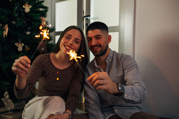 couple holding christmas sprinklers, while decorating christmas tree at home