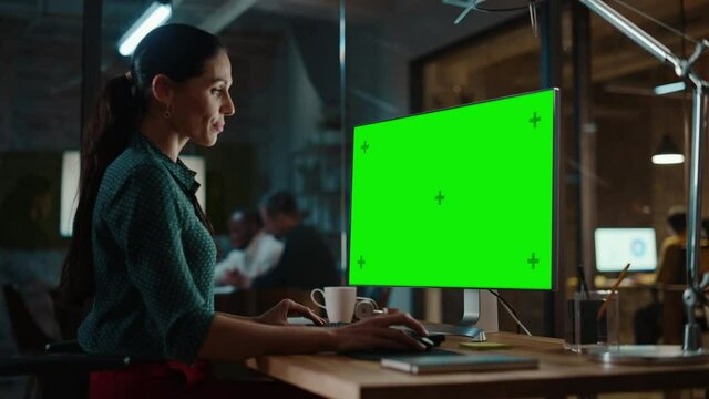 Middle Aged Multiethnic Specialist Working on Desktop Computer with Green Screen Mock Up Display in Busy Creative Office. Beautiful Diverse Female Manager in Green Polka Dot Blouse.
