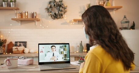 Over shoulder view of Caucasian woman in medical mask having online video conference on laptop with male doctor in quarantine in christmas period. Video consultation on computer on holidays