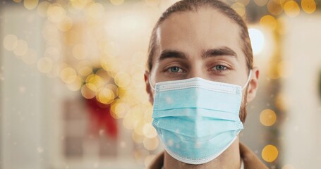 Close up of joyful Caucasian young handsome man in medical mask standing on decorated christmassy street in city looking at camera and smiling with happy face on Xmas Eve Winter holidays in quarantine