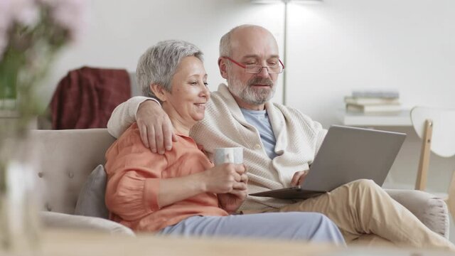 Medium shot of senior Asian woman and Caucasian bearded man sitting in hug on couch in living room, using laptop and talking. Woman holding tea cup