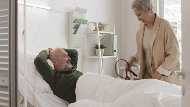 Medium shot of aged bearded Caucasian man with oxygen tube in his nose lying in bed in hospital ward and greeting his attractive senior wife coming to visit him