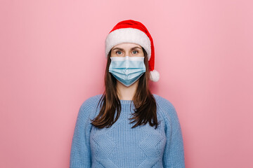 Portrait of young caucasian woman with medical face mask and red Santa hat, looking at camera,...