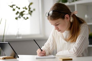 A young girl sit at the table at home and writes in a notebook, studies foreign languages, online...