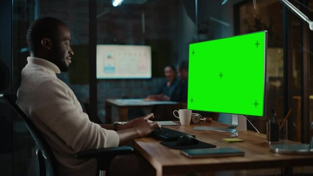 Handsome Black African American Specialist Working on Desktop Computer with Green Screen Mock Up Display in a Busy Creative Office. Male Manager is Wearing a Casual White Turtle Neck Sweater.