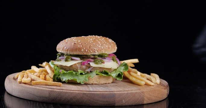 Delicious burger suddenly appears in dark frame on wooden board. Male hands in latex gloves chef put push on black table set of fast food chisburger and french fries fried potatoes in restaurant bar