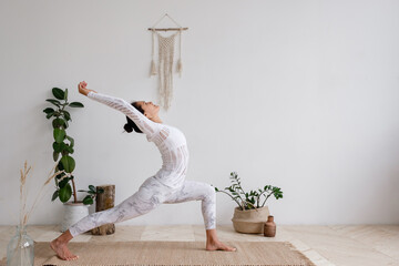 Side view of a slim pretty positive young brunette woman doing a virabhadrasana standing on the mat on the floor surrounded by houseplants on a white wall. Advertising space. Yoga and pilates