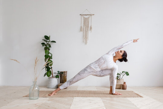 Side view of slim pretty positive young brunette woman doing Utthita parsvakonasana exercise, Extended Side Angle pose, on mat on floor surrounded by houseplants on white wall. Advertising space.