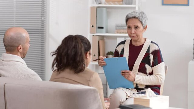 Senior Asian grey-haired female therapist holding clipboard with papers sitting on chair, talking to unrecognizable couple at appointment, woman getting emotional, taking paper tissue