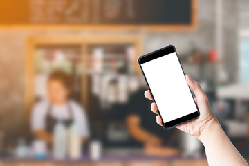 Shopping online concept. Hand holding black smartphone and white blank screen in blurry at coffee shop background. Mock up shopping.