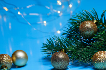 Christmas background. Golden balls and a branch of artificial spruce on a light blue background with garland lights. Side view, horizontal, close-up, free space at the top.