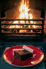 Traditional christmas cake on a red plate with cheese, in front of a roaring log fire