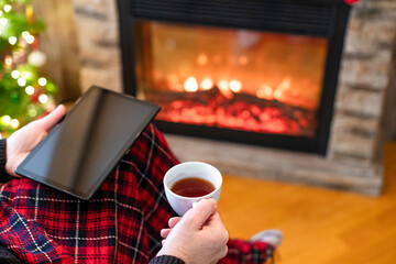 Christmas. Man in santa claus hat with tablet and cup of tea sitting near christmas tree and fireplace. Concept of online service in holidays. Back view.