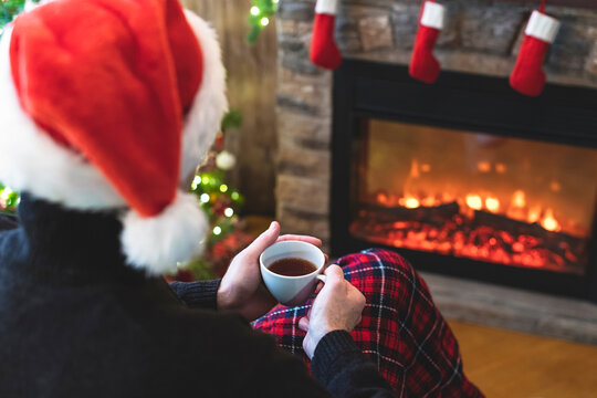One divorce man with cup coffee, tea in santa claus hat sitting and warming at winter evening near fireplace flame, covered christmas plaid
