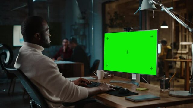 Handsome Black African American Project Manager is Making a Video Call on Desktop Computer with Green Screen Mock Up Display in a Busy Creative Office. Male Specialist is Wearing Turtle Neck Sweater.