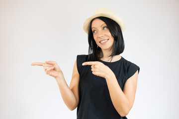 Young brunette woman with blue eyes wearing straw hat casual black t-shirt standing over background smiling and looking at the camera pointing with two hands and fingers to the side.