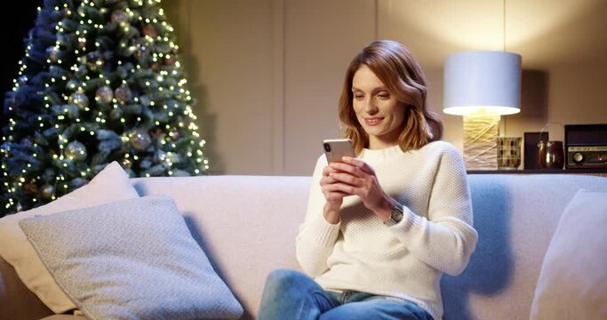 Joyful Caucasian beautiful woman in good mood sitting in cozy room near glowing xmas tree typing on smartphone with happy face. Happy New Year. Merry Christmas concept. Winter holidays