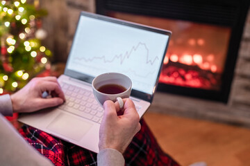 Man freelancer working on laptop with graphs and charts on screen sitting near christmas tree and fireplace with cup of tea.