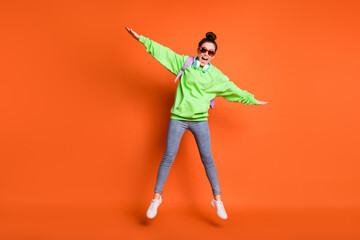 Fototapeta na wymiar Photo portrait full body view of excited schoolgirl jumping up making wings with hands isolated on vivid orange colored background