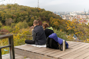 Fototapeta premium A sweet lovely couple enjoys time together on the observation deck with thermos, looking at the city and autumn nature