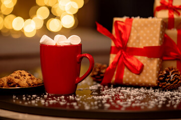 Obraz na płótnie Canvas Red mug of hot cocoa with marshmallows, pine cones, gifts and cookies with chocolate on a snowy table against a background with beautiful Christmas bokeh lights. Blurred background.