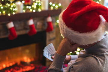 Sad man in a santa claus hat thinking counting american dollars sitting near christmas tree and fireplace. Spending money on gift at christmas time. concept.