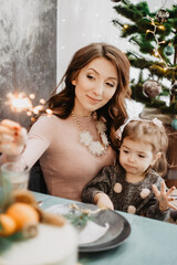 Young family with a daughter in festive outfits at a served table with candles, garlands, sparklers and a cake near the Christmas tree on New Years Eve