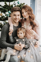 Young family with a daughter in festive outfits with a garland near the Christmas tree on New Years Eve