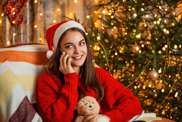 Attractive teenager girl sitting in the armchair, holding teddy bear and talking over the phone. Happy family time, festive atmosphere, warm cozy interior.