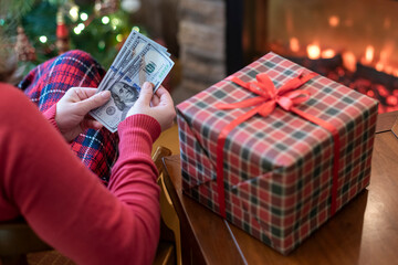 Woman counting american dollars planning sitting near christmas tree and fireplace and packing gift box. Spending money at christmas time. Сoncept.