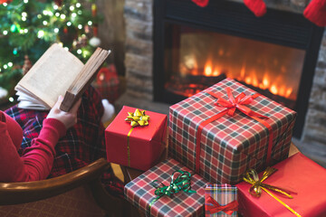 Fototapeta na wymiar Alone woman covered plaid reading book sitting and relaxation on armchair near fareplace and christmas tree after finishing pakking gift boxes for family.