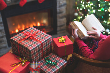 Alone woman covered plaid reading book sitting and relaxation on armchair near fareplace and christmas tree after finishing pakking gift boxes for family.