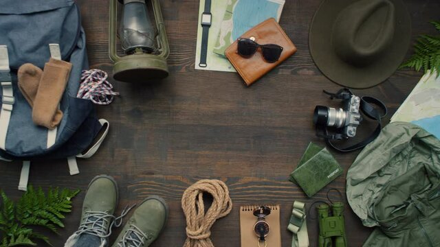 Top view zoom out flat lay of passports, boots, rope, compass, binoculars, raincoat, photo camera, hat, maps, backpack, lantern and warm socks lying on wooden table decorated with fern