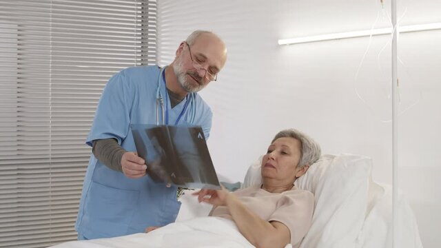 Medium shot of ill old Asian woman lying in bed in hospital room and elderly Caucasian doctor wearing blue uniform standing nearby and showing x-ray image to her