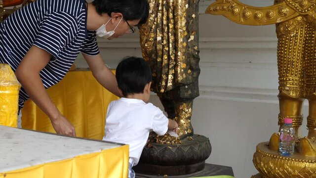 4K Buddhist asian child boy and dad apply gold leaf to buddha statue, son and father having activity together, making merit in local temple in countryside. Happy family activity in holiday.