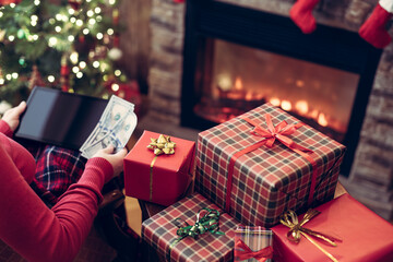 Naklejka na ściany i meble Christmas. Woman in sweater with tablet and american dollars searching gift sitting at table near fireplace and christmas tree. Concept