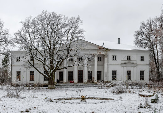 An old abandoned white manor in winter in the snow. A large tree next to an antique building. Beautiful ancient architecture with columns.
