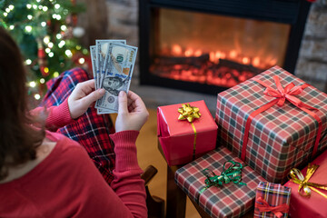 Woman counting american dollars planning sitting near christmas tree and fireplace and packing gift...