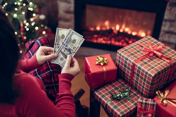 Woman counting american dollars planning sitting near christmas tree and fireplace and packing gift...