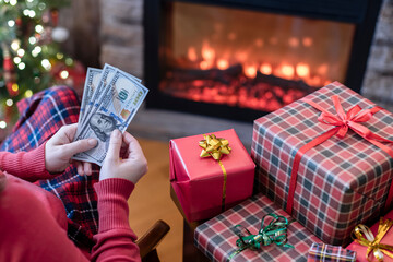 Obraz na płótnie Canvas Woman counting american dollars planning sitting near christmas tree and fireplace and packing gift box. Spending money at christmas time. Сoncept.