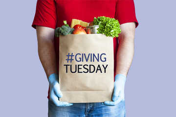 Volunteer in medical gloves holds paper grocery bag with inscription Giving tuesday on blue background. Charity, help and donation concept.
