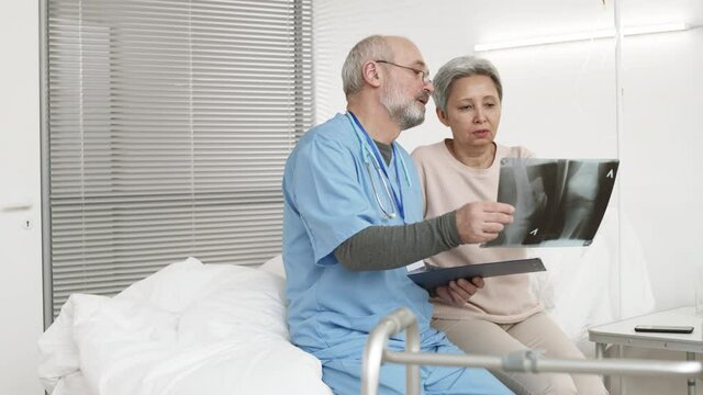 Medium shot of senior Asian woman and aged Caucasian doctor wearing blue uniform sitting on bed in hospital ward and analyzing x-ray picture