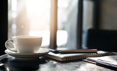 Close-up white coffee cup with notebook, pen and mobile on table with sunlight winter on the morning