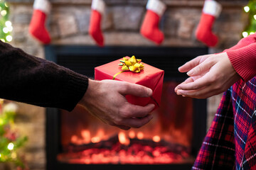 Man giving Christmas and New Year Gift box to woman in front of christmas tree, fireplace with flame. Xmas concept.