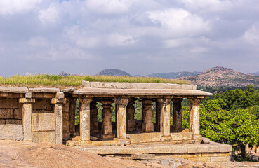Obraz premium Hampi, Karnataka, India - November 4, 2013: Virupaksha Temple complex. Moola brown stone gallery ruin with grass growing on top under light blue cloudscape and green foliage around.