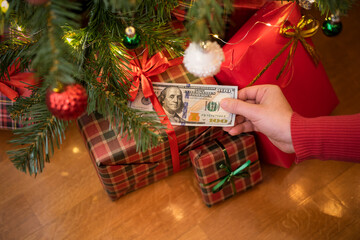 Woman putting american dollars under red bow of huge gift box lying under christmas tree. Concept