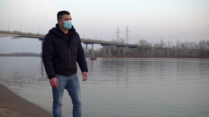 A man in a mask on his face walks along the river bank